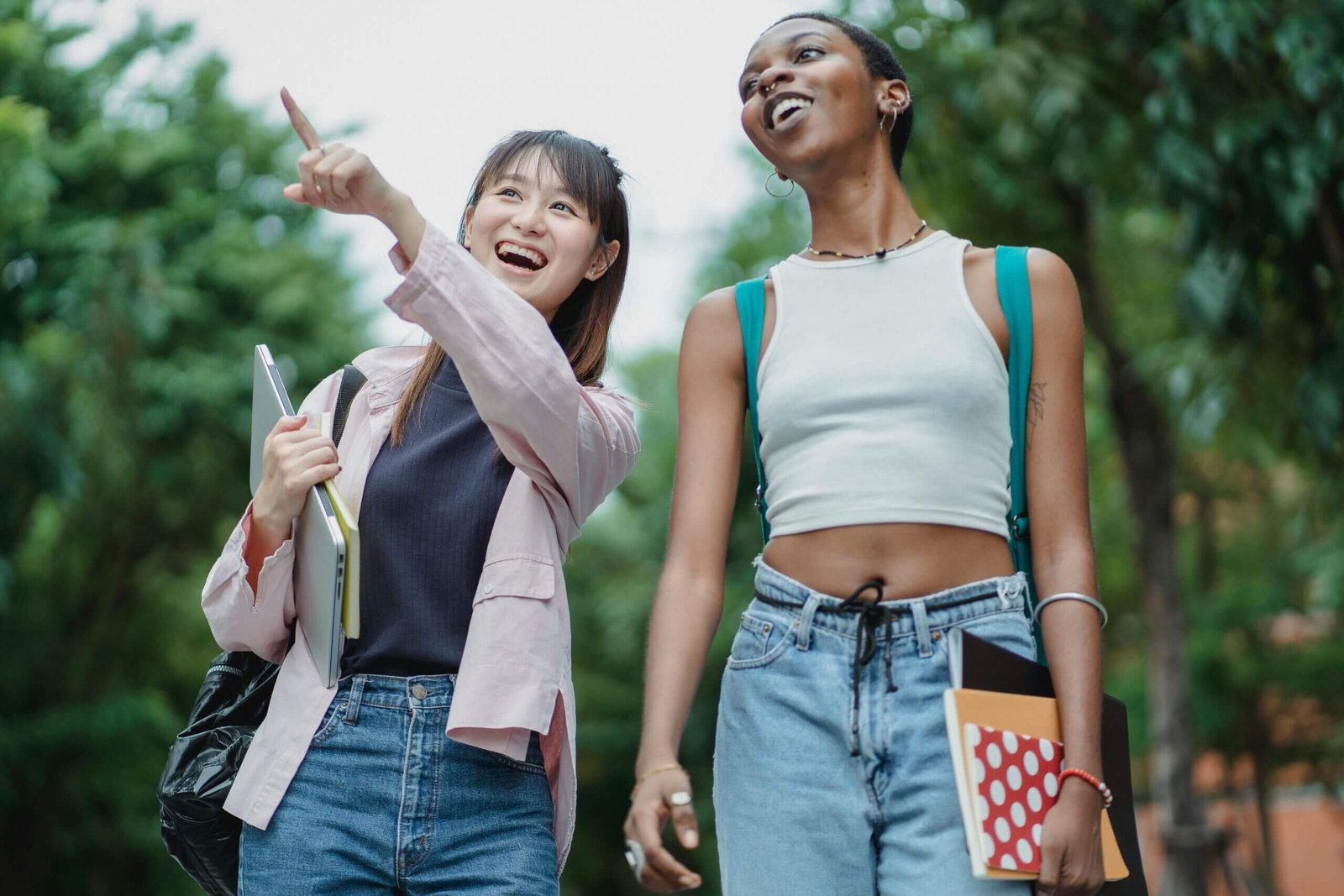 Cheerful Asian schoolgirl with laptop and copybooks pointing away while strolling together with smiling African American friend in lush summer park